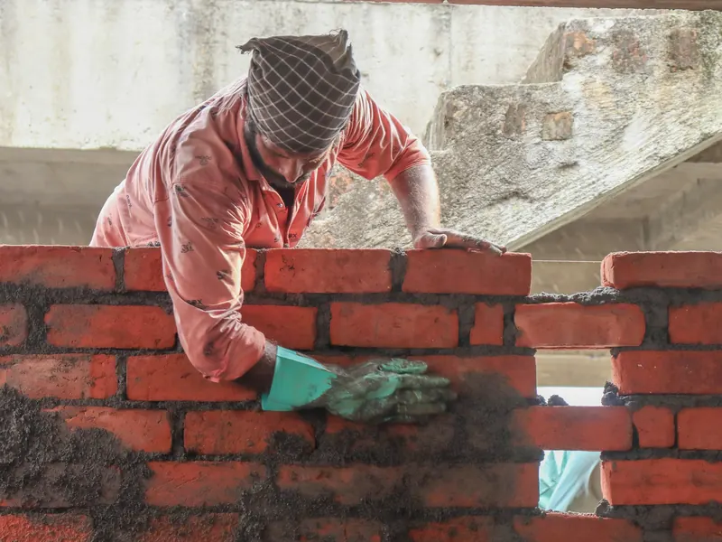 Mason laying a course of red kiln bricks on a rising wall, spreading mortar with a trowel.