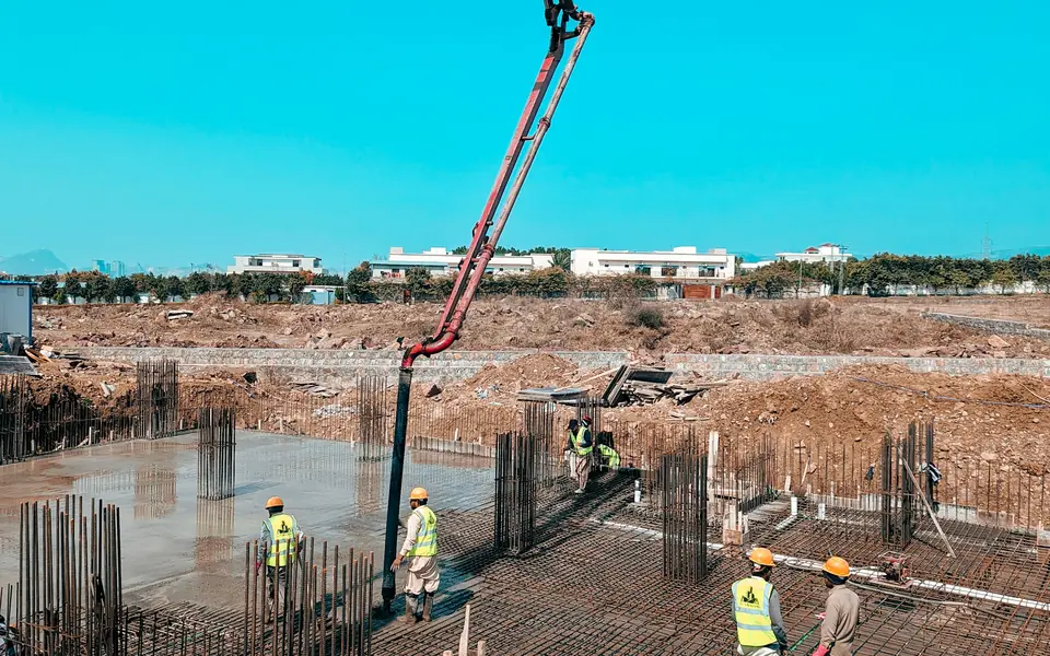 Concrete boom pump discharging into a reinforced raft foundation while workers in high-visibility vests spread and level the pour.